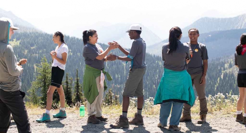 a group of outward bound students stand on an overlook while two play patty cake with their hands
